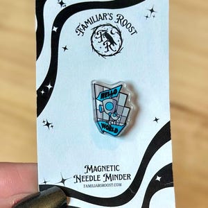 May include: A magnetic needle minder with a clear, geometric design featuring the words "Hello World" and a small robot illustration. The minder is attached to a black and white patterned card with the text "Familiar's Roost".