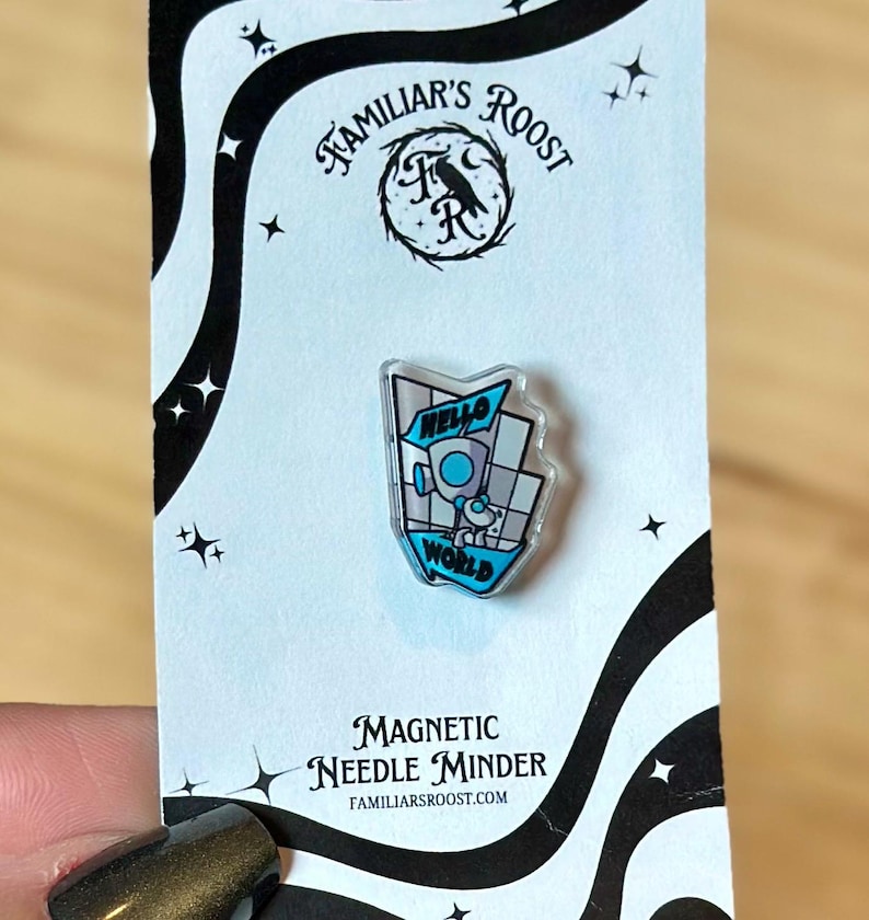 May include: A magnetic needle minder with a clear, geometric design featuring the words "Hello World" and a small robot illustration. The minder is attached to a black and white patterned card with the text "Familiar's Roost".