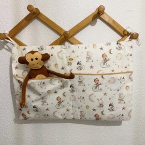 Può includere: Organizzatore appendibile in tessuto bianco con due tasche. L'organizzatore presenta un motivo fantasioso di animali che vanno in bicicletta, stelle e lune. Un peluche di scimmia marrone si trova nella tasca superiore.