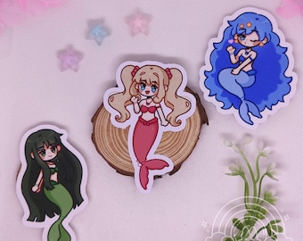 Op de afbeelding: Drie zeemeermin stickers met verschillende haar- en staartkleuren. Eén zeemeermin heeft blonde staartjes en een rode staart, een andere heeft blauw haar en een blauwe staart, en de derde heeft donkergroen haar en een groene staart. De stickers liggen op een houten schijf.