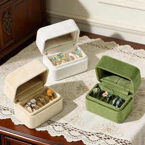 May include: Three rectangular velvet ring boxes in white, beige, and olive green, each featuring a hinged lid and interior slots holding various rings with colorful gemstones and metallic bands, arranged on a white lace-trimmed fabric surface.