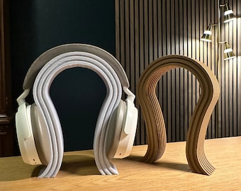 May include: Two headphone stands, one made of wood and one made of white plastic. The wooden stand is a simple, curved design, while the plastic stand is more elaborate, with multiple layers.