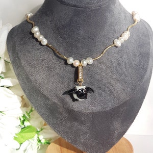 May include: A gold chain necklace with white pearls and a black and white sheep charm. The necklace is displayed on a grey velvet bust.