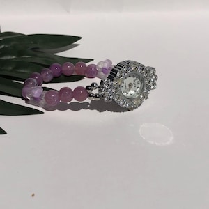May include: A beaded bracelet watch with a silver-colored watch face. The watch face is surrounded by clear crystals. The bracelet is made of pink and purple beads. The watch is on a white surface.