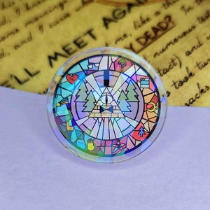 May include: A round, clear glass pin with a colorful stained glass design featuring a triangle with an eye inside, surrounded by symbols and a pine tree design. The text "WE'LL MEET AGAIN" is visible in the background.