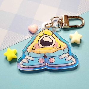 May include: A yellow and blue iridescent keychain featuring a cartoon character with one large eye, wearing blue sneakers with baseballs on them.