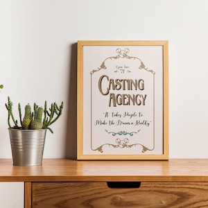 Pode incluir: Uma impressão emoldurada com as palavras "Casting Agency" em estilo vintage, com enfeites decorativos. A impressão está sobre uma superfície de madeira, com um vaso branco e um cacto em vaso por perto.
