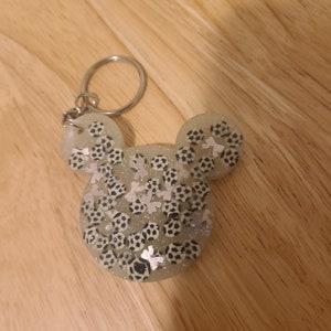 May include: A white resin keychain shaped like Mickey Mouse's head, filled with black and white footballs and silver glitter.