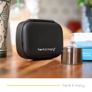 Könnte beinhalten: Eine schwarze, rechteckige Tragetasche mit Griff, mit dem weißen Text "herb & mary". Ein kleiner, silberner Metallbehälter ist ebenfalls sichtbar. Die Artikel befinden sich auf einer Holzoberfläche.