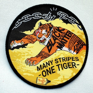 May include: A black and orange tiger with the words "Black Lives Matter" and "Many Stripes - One Tiger" on a yellow background. The tiger is wearing a chain around its neck and has the words "Queer Liberation" and "Abolish Now" written on its body.