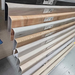 May include: A display of various wood-look baseboards in shades of gray, brown, and beige. The baseboards have a curved top edge and are stacked, showcasing different colors and textures. Each piece has a label attached.