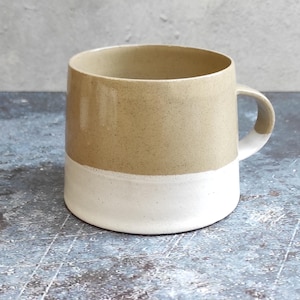 May include: A two-tone ceramic mug. The upper half is beige, the lower half is white. The mug has a rounded shape with a handle on the right. It sits on a blue and grey surface.