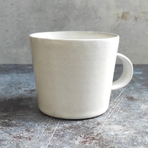 May include: A speckled, off-white ceramic mug with a rounded body and a curved handle. The mug has a simple, minimalist design and appears to be handmade. The mug is sitting on a textured surface.