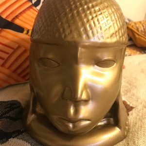 Puede incluir: Un macetero de cerámica de color dorado con forma de cabeza humana. El macetero tiene una superficie texturizada y un diseño simple y minimalista.