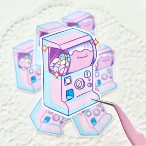 May include: A pink and blue gumball machine with a cartoon frog face on the front. The machine is filled with colorful gumballs and has a sparkling effect.