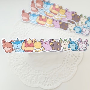 May include: A row of colorful cartoon stickers featuring various Pokemon characters, including Vaporeon, Flareon, Jolteon, and others. The stickers are arranged in a line on a white surface.