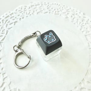 May include: A black keycap keychain with a cartoon character design in blue and white. The keycap is attached to a silver metal spring and a keyring. The keychain is on a white, textured surface.