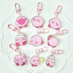 Puede incluir: Una colección de nueve llaveros de acrílico transparente con varios personajes de dibujos animados de color rosa con diferentes expresiones y accesorios, cada uno unido a un mosquetón rosa claro. Los diseños incluyen personajes con sombreros, sosteniendo lana o comiendo pastel.