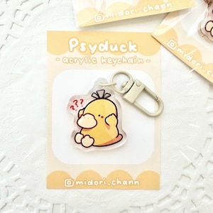 Puede incluir: Un llavero de acrílico con el personaje de Psyduck, un Pokémon amarillo. El llavero tiene un pequeño signo de interrogación blanco sobre la cabeza del personaje. El llavero está unido a un anillo y un cierre de metal plateado.