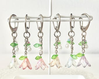 Op de afbeelding: Zes sleutelhangers met bloemvormige bedels. Elke sleutelhanger heeft een zilverkleurige sluiting en ketting, met een doorschijnende bloembedel in verschillende kleuren: helder, roze en paars. De bedels zijn versierd met groene bladeren en witte parel kralen.