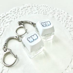 May include: Two keychains with white keycaps featuring a blue cloud and bear design. The keycaps are mounted on clear, rectangular blocks. Each keychain has a silver-colored ring and a coiled metal attachment.