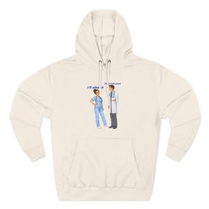 May include: Cream-colored hoodie with a front pocket and drawstring hood. The graphic depicts a nurse and a doctor with the text "I'll allow it" and "Here's the plan". The illustration is in blue and the hoodie is made of a soft material.