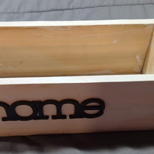 May include: A rectangular wooden box with the word "home" in black lettering on the front. The box is made of light-colored wood and has a simple, clean design. The box is empty.
