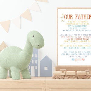 May include: A colorful printable wall art print with the Lord's Prayer, "Our Father", in a rainbow font. The print is framed and hanging on a wall in a nursery with a green dinosaur toy and a wooden train set.