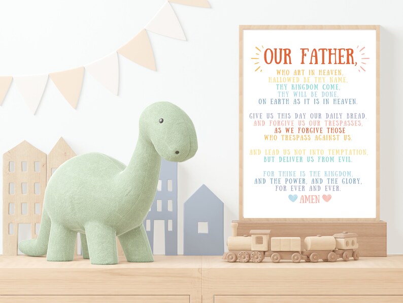 May include: A colorful printable wall art print with the Lord's Prayer, "Our Father", in a rainbow font. The print is framed and hanging on a wall in a nursery with a green dinosaur toy and a wooden train set.