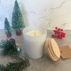 May include: A white candle in a glass jar with a wooden lid. The candle is surrounded by Christmas decorations, including small green trees, red berries, and pine branches. The candle has a neutral, minimalist aesthetic.