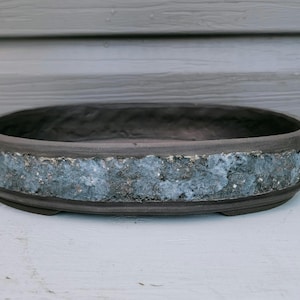 May include: An oval-shaped ceramic bonsai pot with a dark brown, smooth interior and rim. The exterior side features a wide, textured band with a mottled, rough finish in shades of grey, blue, and white, contrasting with the dark brown base.