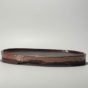 Peut inclure: Un pot à bonsaï en céramique marron, de forme rectangulaire aux bords arrondis. L'intérieur est brun foncé et l'extérieur est brun rougeâtre texturé avec un éclat métallique. Conçu pour les bonsaïs, environ 30 cm de long.