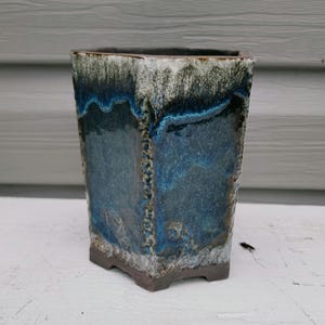 May include: A hexagonal ceramic planter featuring a reactive glaze with deep blue, teal, and white tones. The surface displays a textured, dripped finish that transitions from a dark base to a lighter rim, with visible vertical seams at the corners.