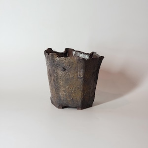 May include: A textured, brown ceramic planter with an irregular rim. The planter has a geometric, multi-sided shape and sits on small feet. The surface has a rough, aged appearance, suggesting a handcrafted or rustic aesthetic.