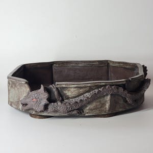 May include: A rectangular, ceramic planter with a dragon design. The planter is a muted grey-brown colour with a dragon sculpted on the side. The dragon has red eyes and detailed scales. The planter is approximately 30 cm long.
