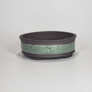May include: A dark brown ceramic bonsai pot with a light green textured band around the middle. The oval-shaped pot sits on three small feet. The pot is designed for growing small trees and plants.