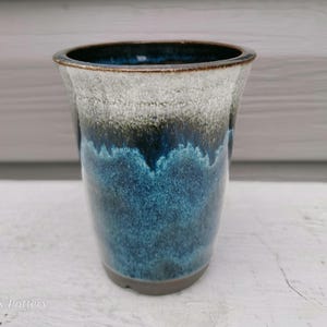 Peut inclure: Un gobelet en céramique doté d'un émail réactif avec un bord blanc qui se transforme en une bande noire profonde, suivie d'une base bleu vif. Le texte MK Pottery est imprimé dans le coin inférieur gauche de l'image en police blanche.