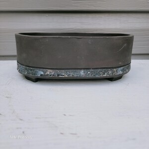 May include: An oval-shaped ceramic bonsai pot with a matte dark charcoal finish on the upper portion and a textured, mottled blue and grey glaze along the base. The vessel features a smooth rim and is supported by small, integrated feet.