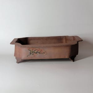 May include: A rectangular, brown bonsai pot with a decorative tree branch and cloud design. The pot has a textured surface, four small feet, and a slightly raised rim. The pot is made of ceramic.
