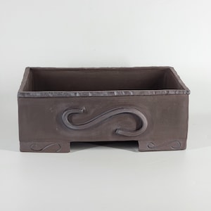 Puede incluir: Una jardinera rectangular de cerámica marrón con un diseño de voluta en relieve en la parte delantera. La jardinera tiene un borde texturizado y pies decorativos. El interior está vacío, listo para plantas u otros elementos decorativos.