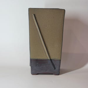 Könnte beinhalten: Eine rechteckige Keramikvase mit einem beigefarbenen oberen Bereich und einem dunkelgrauen unteren Bereich. Eine diagonale Linie verläuft über die Vorderseite. Die Vase hat ein minimalistisches Design und eignet sich für die Inneneinrichtung.