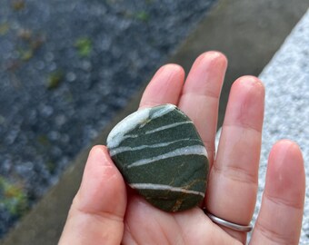 May include: A small, smooth, green stone with white horizontal stripes. The stone is held in an open hand, showcasing its natural patterns and colors. The stone's surface appears slightly wet, suggesting it was recently found.