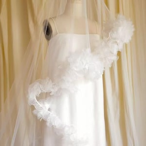 Puede incluir: Un vestido de novia blanco con un velo transparente y un ribete decorativo con volantes. El vestido se exhibe en un maniquí sobre un fondo suave de color crema. El velo cae por la parte delantera del vestido.