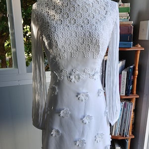 Op de afbeelding: Witte jurk met lange mouwen en bloemenkanten details. Het lijfje en de mouwen hebben ingewikkelde kanten patronen, terwijl de rok is versierd met delicate bloemenapplicaties. Een klassiek en elegant ontwerp.