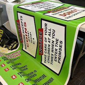 May include: Green and white printed signs with black text. One sign reads "NOTICE MACHINE ACCEPTS COINS" with coin denominations. Another sign says "TAKE CARE OF YOUR CHILDREN AT ALL TIMES WHEN YOU ENTER THE PRIMSES."
