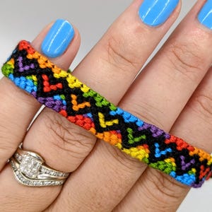 Peut inclure: Un bracelet d'amitié coloré avec un motif en chevrons aux couleurs de l'arc-en-ciel : rouge, orange, jaune, vert, bleu et violet. Le bracelet est présenté sur un fond blanc, tenu par une main avec du vernis à ongles bleu et une bague en diamant.