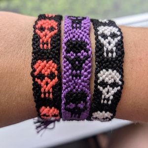 May include: Three friendship bracelets with skull designs. The bracelets are black, purple, and orange with white accents.