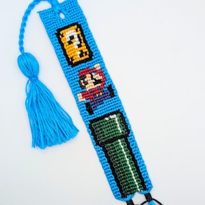 Könnte beinhalten: Ein blaues und grünes Freundschaftsarmband mit einem Pixelbild von Mario, einem Fragezeichenblock und einem grünen Rohr. Das Armband hat an einem Ende einen blauen Quasten und an dem anderen Ende zwei blaue Quasten.