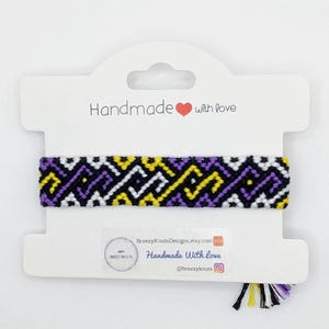 May include: A handmade friendship bracelet with a geometric pattern in black, white, purple, and yellow. The bracelet is presented on a white card with the text "Handmade with love". The card also displays the Etsy shop name and social media handle.