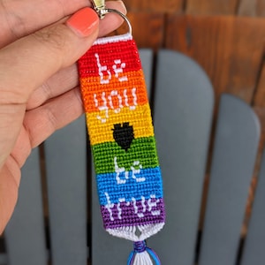 May include: A rainbow-colored keychain with a black heart and the words "Be You Be True" woven into the fabric. The keychain has a silver metal clasp.